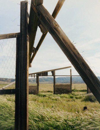 Dilapidated wooden structure in a grassy field under a light blue sky. Weathered wood and chicken wire fence with a rural backdrop.