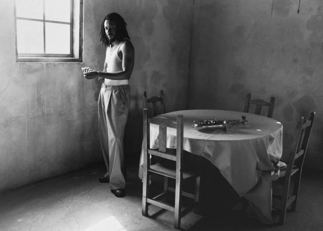 Black and white shot of a slender man with dreadlocks standing near a window in a textured room with a saxophone on a table.