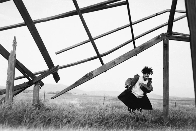 Black and white portrait of a man in a coat standing in a grassy field near a dilapidated wooden structure, rural landscape. Documentary style.
