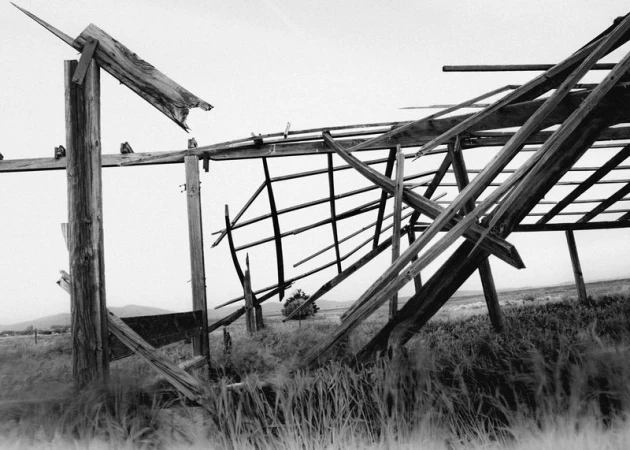 Monochrome image of a dilapidated barn structure in a grassy field with a distant mountain range, creating a stark, abandoned scene.