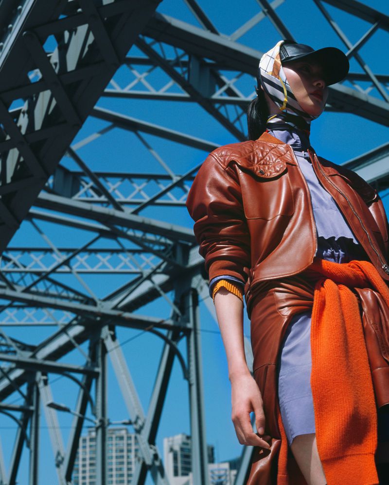 Woman in brown leather jacket, skirt, headscarf, and baseball cap stands under a steel bridge on a sunny day with a bright blue sky.