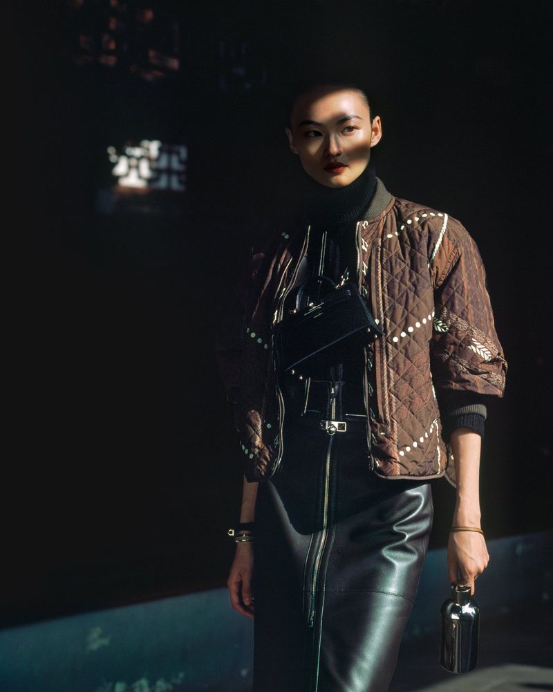 An Asian woman with a shaved head poses in dark studio light, wearing a quilted brown jacket and leather skirt, holding a small metal object.