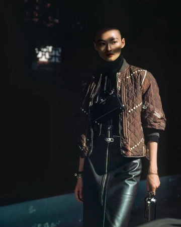 An Asian woman with a shaved head poses in dark studio light, wearing a quilted brown jacket and leather skirt, holding a small metal object.