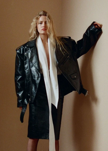 A blonde woman poses in a black leather jacket and skirt, complemented by a flowing white scarf against a neutral backdrop.