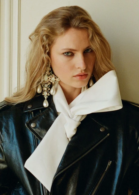 Close-up studio shot of a blond woman wearing a black leather jacket with a white scarf, statement earrings, and natural makeup.