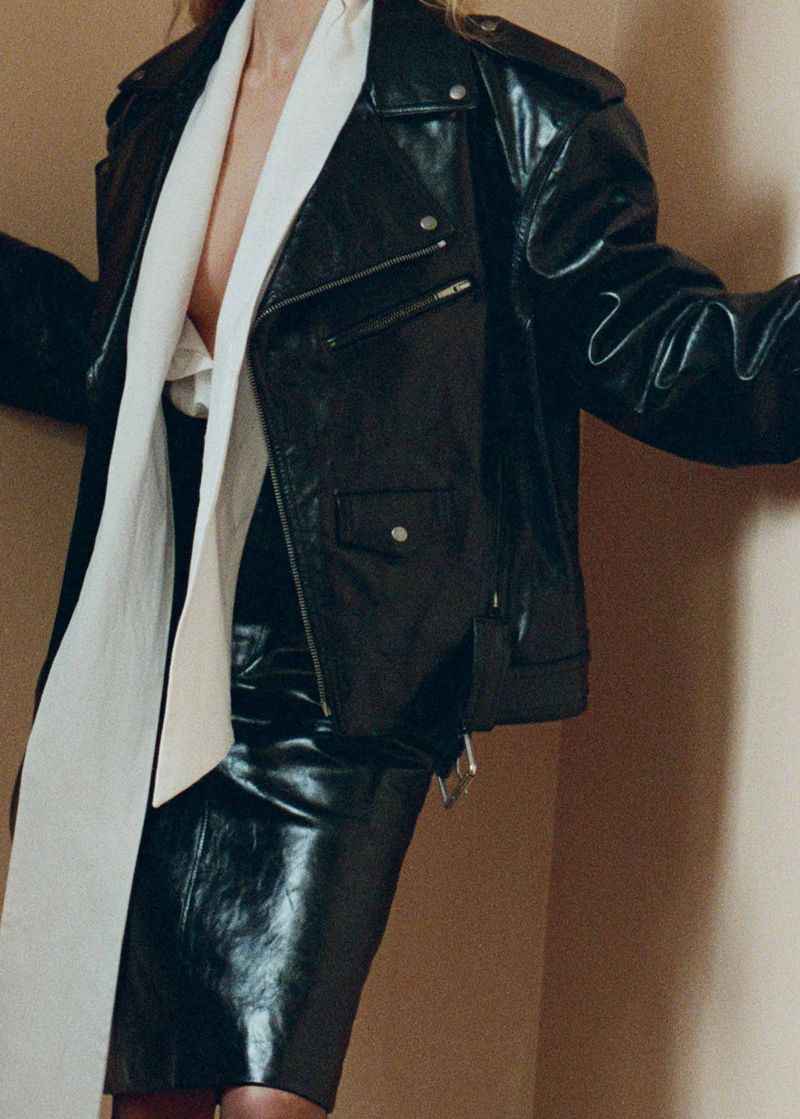 Woman in shiny black leather jacket and skirt with white scarf standing indoors against a neutral-color wall in a fashion editorial.