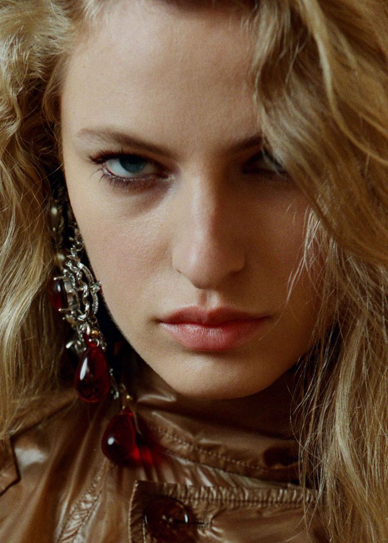 Close-up portrait of a young fair-skinned woman with blonde curly hair wearing large drop earrings and a golden-brown jacket.