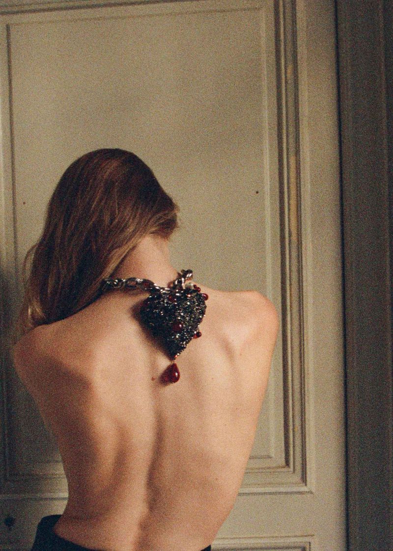 Fashion portrait of a woman's back and shoulders, featuring a large, ornate heart-shaped pendant necklace with a red drop.