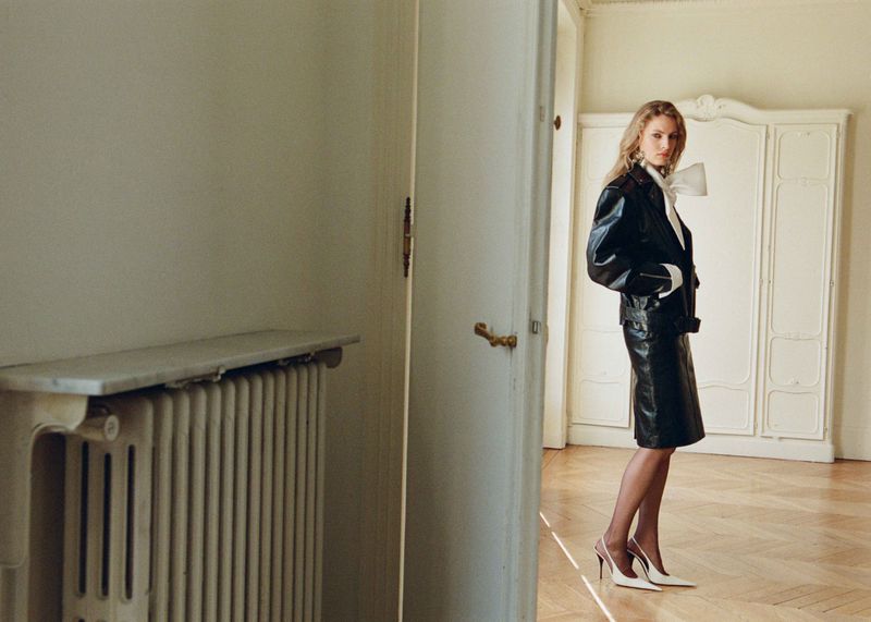 A blonde woman in a black skirt and jacket stands in an old-fashioned room with parquet floors, next to a white door and radiator.