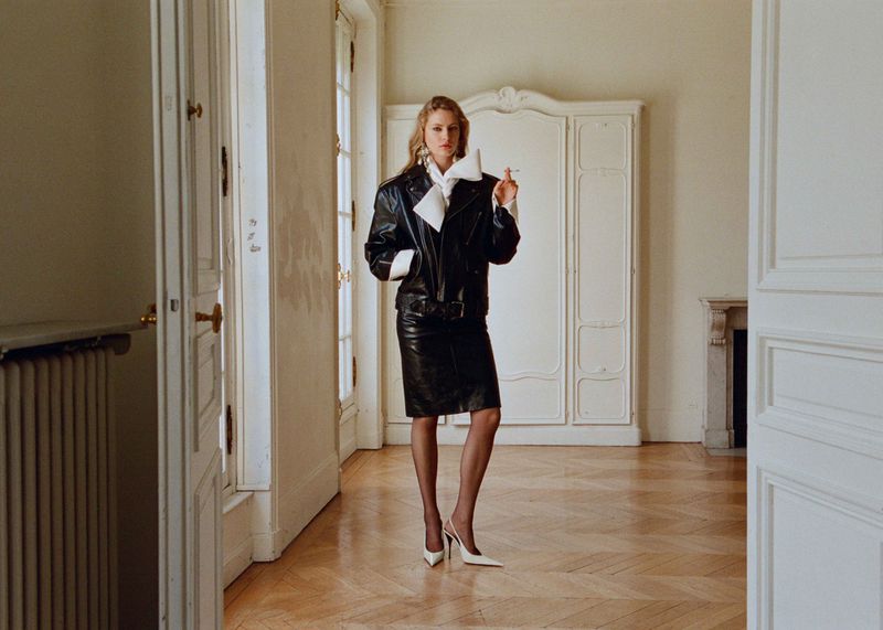 Blonde woman in a black leather jacket and skirt stands in a white room, holding a cigarette with a wardrobe in the background.