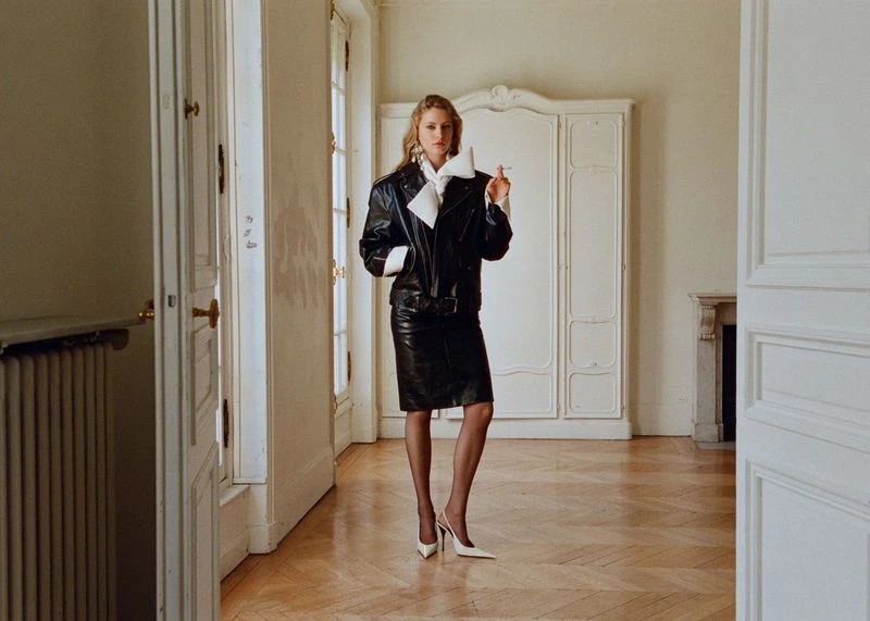 Blonde woman in a black leather jacket and skirt stands in a white room, holding a cigarette with a wardrobe in the background.