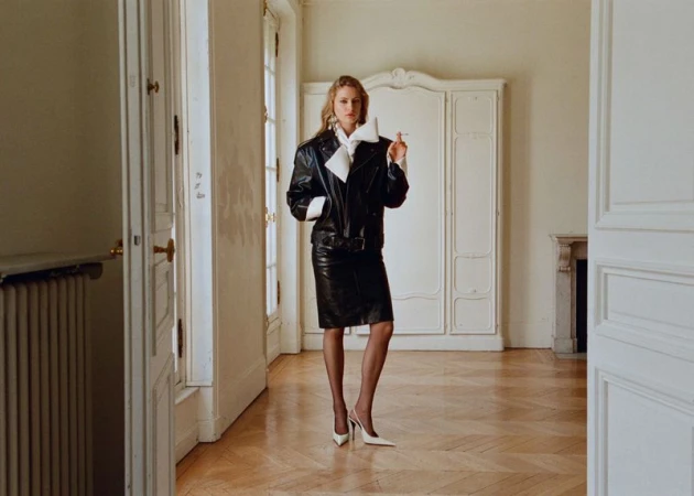 Blonde woman in a black leather jacket and skirt stands in a white room, holding a cigarette with a wardrobe in the background.
