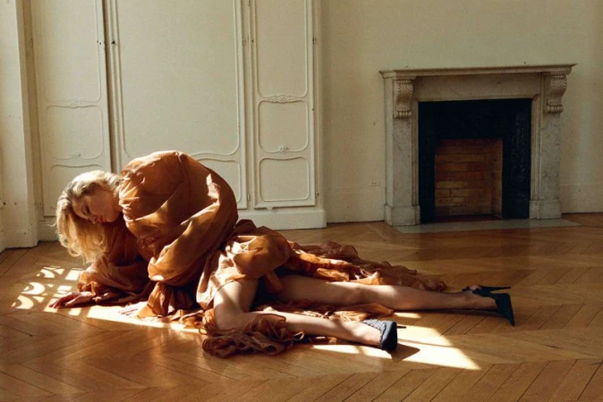 Blonde woman in flowing gown and black heels lies on hardwood floor with sunlight in empty room with paneled walls and fireplace.