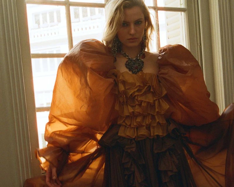 Fashion shot of a woman with blonde hair wearing a ruffled, copper-colored gown, large earrings, and a heart necklace in front of a window.
