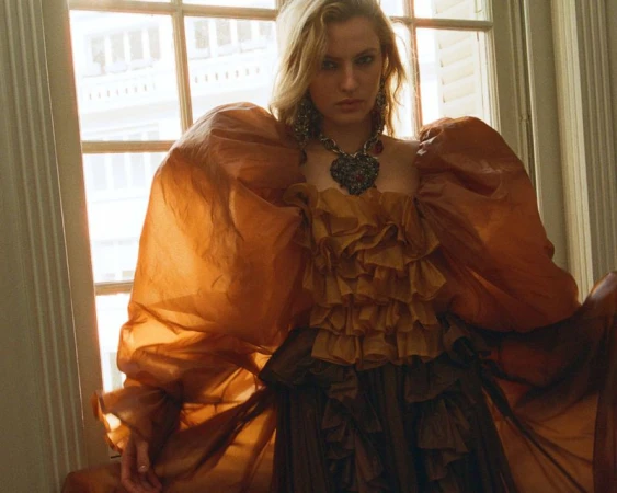 Fashion shot of a woman with blonde hair wearing a ruffled, copper-colored gown, large earrings, and a heart necklace in front of a window.
