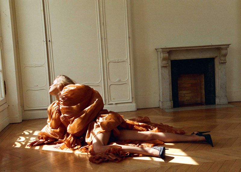 Woman in a flowing copper-colored dress and heels poses on a wood floor in a sunlit interior next to a window and decorative fireplace.