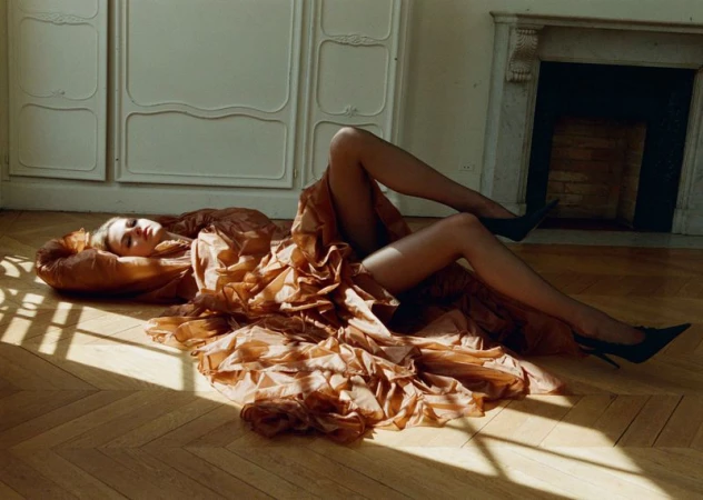 Young adult woman lying on the floor in a ruffled copper dress, black stockings and high heels in a room with wood floors and a fireplace.