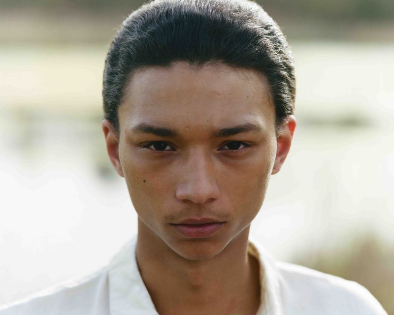 Close-up of a young adult male with dark hair and light skin, wearing a white shirt, looking directly at the camera outdoors.