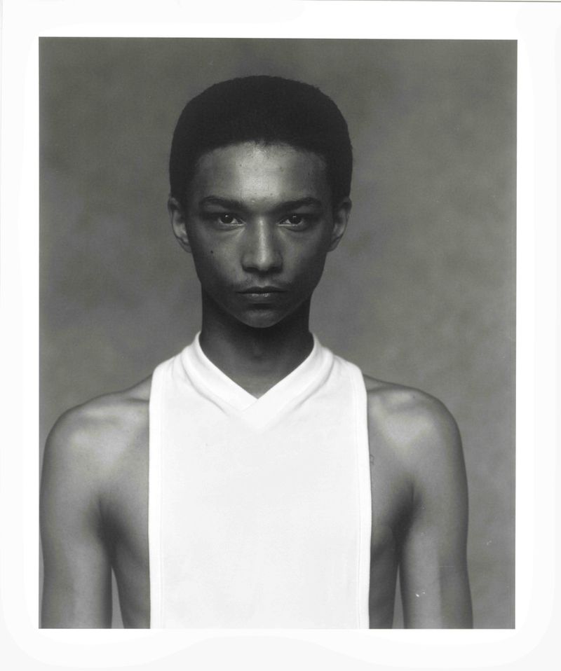 Close-up portrait of a young adult with short hair and a white tank top against a grey background, high-key lighting, neutral expression.