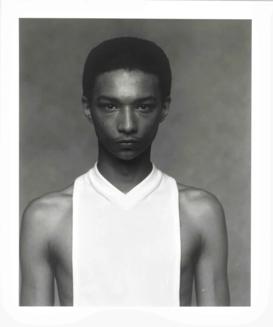 Close-up portrait of a young adult with short hair and a white tank top against a grey background, high-key lighting, neutral expression.