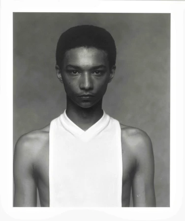 Close-up portrait of a young adult with short hair and a white tank top against a grey background, high-key lighting, neutral expression.