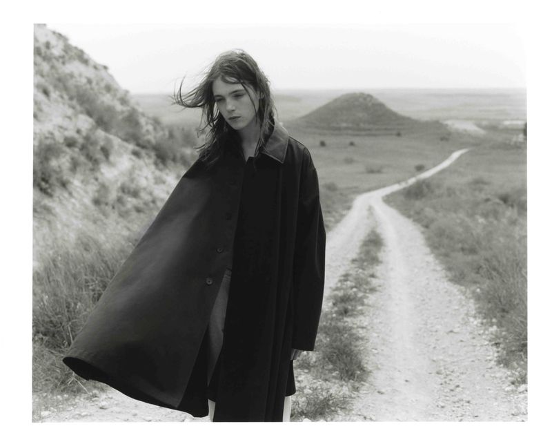 Monochrome outdoor fashion shot: Woman in dark coat walks on dirt path flanked by grassy hills in a desolate landscape, wind blowing.