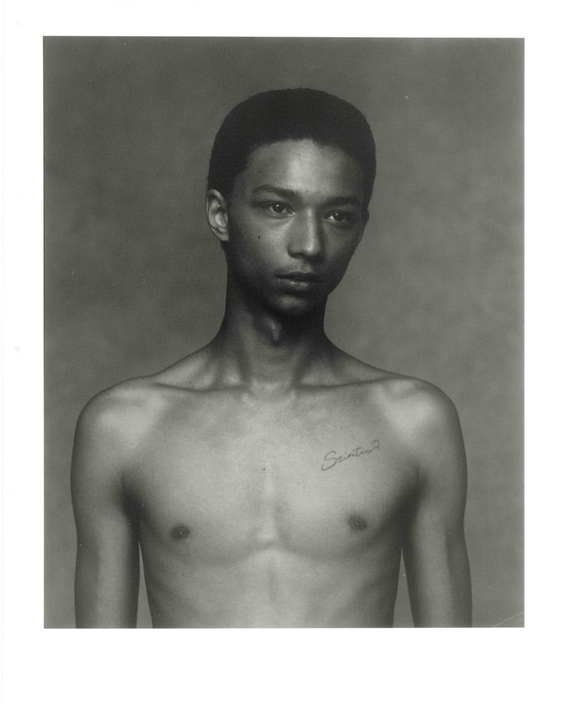 Studio portrait of a shirtless young adult male with a tattoo, standing against a neutral grey background, captured in black and white.