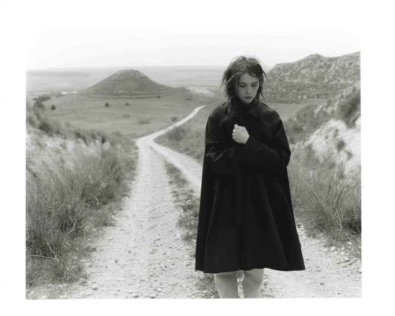 Monochrome shot of a young woman in a dark coat standing on a dirt path in a hilly landscape with fields and a cloudy sky.