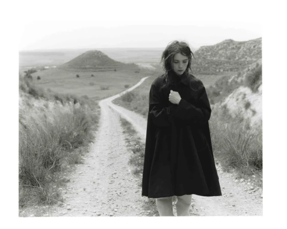 Monochrome shot of a young woman in a dark coat standing on a dirt path in a hilly landscape with fields and a cloudy sky.