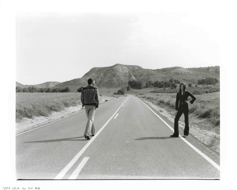 Monochrome landscape with a man walking away on a road and a woman standing on the roadside, backed by rolling hills.