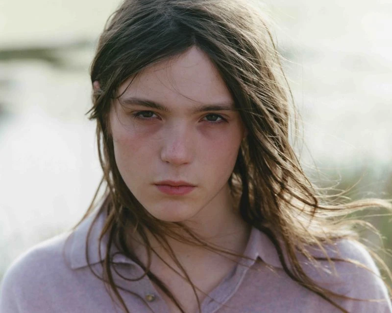 Close-up portrait of a young adult with long, light brown hair and fair skin, wearing a collared shirt, with a blurry, natural background.