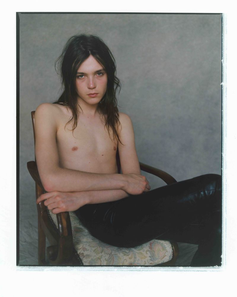 Portrait of a young adult with long hair sitting in a wooden chair, posing shirtless and wearing black leather pants against grey backdrop.
