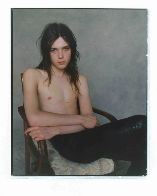 Portrait of a young adult with long hair sitting in a wooden chair, posing shirtless and wearing black leather pants against grey backdrop.