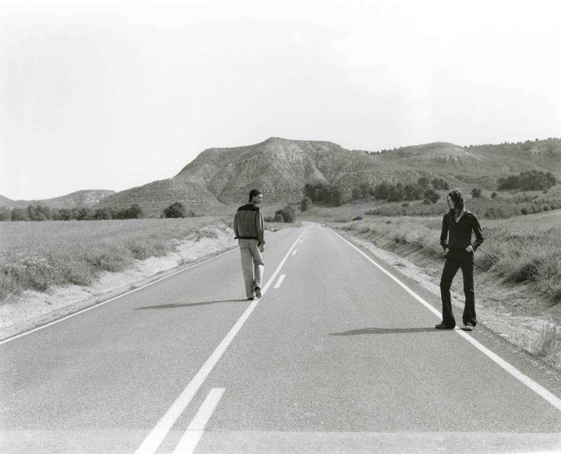 Black and white photograph of two people standing on an empty road in a rural landscape, with rolling hills in the background under a clear sky.
