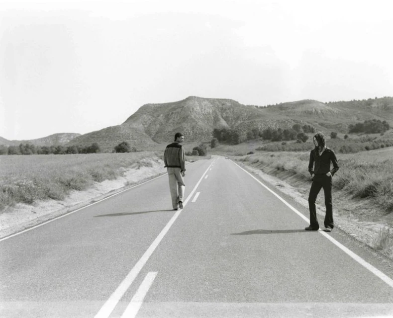 Black and white photograph of two people standing on an empty road in a rural landscape, with rolling hills in the background under a clear sky.