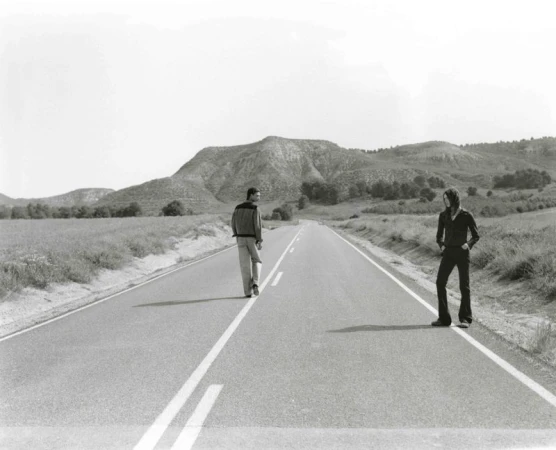 Black and white photograph of two people standing on an empty road in a rural landscape, with rolling hills in the background under a clear sky.