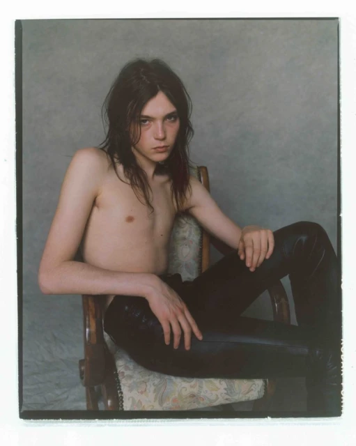 Young adult with long dark hair poses in a chair wearing shiny black pants against a mottled grey background in a studio setting.