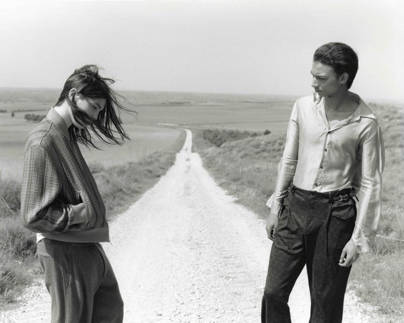 Two people stand on a dirt road in a rural landscape on a cloudy day in a black-and-white fashion photograph.