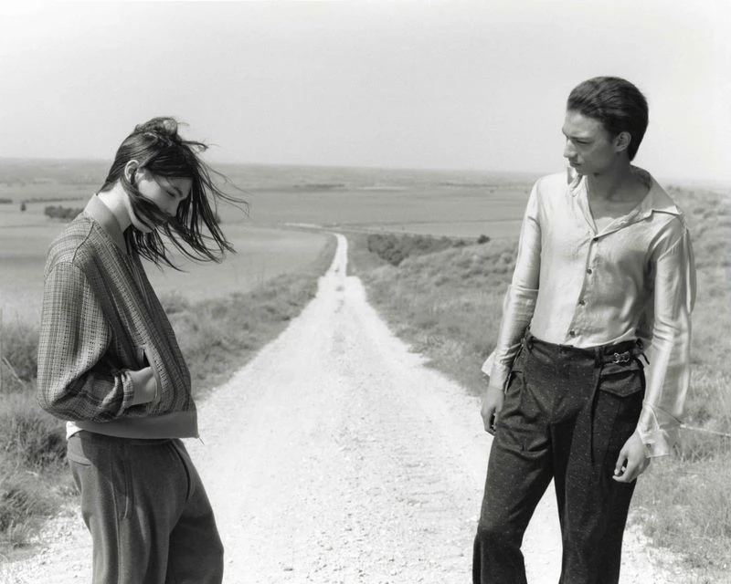 Two people stand on a dirt road in a rural landscape on a cloudy day in a black-and-white fashion photograph.