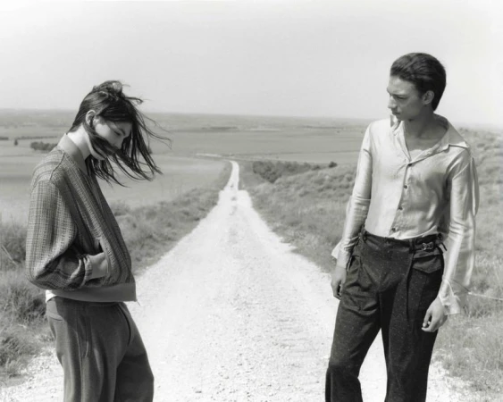 Two people stand on a dirt road in a rural landscape on a cloudy day in a black-and-white fashion photograph.