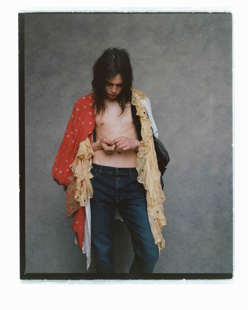 Young adult with long dark hair stands indoors wearing blue jeans and a layered red floral and cream-colored ruffled top, looking down.