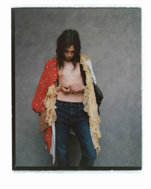 Young adult with long dark hair stands indoors wearing blue jeans and a layered red floral and cream-colored ruffled top, looking down.