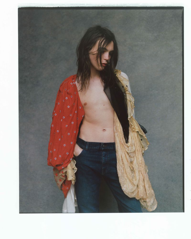 Young adult with long brown hair, wearing jeans and draped in sheer fabric and red patterned shirt against a grey backdrop.
