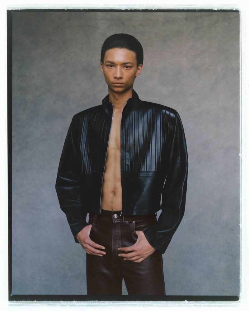 A model poses in a studio with a black leather jacket and brown leather pants in a medium shot with a neutral expression.