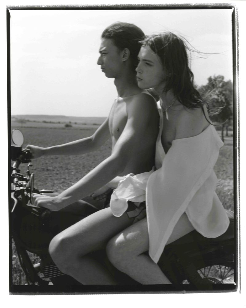 Black and white photograph of a young couple riding a motorcycle through a rural landscape on a sunny day, the woman wearing a white garment.