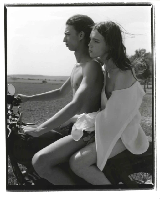 Black and white photograph of a young couple riding a motorcycle through a rural landscape on a sunny day, the woman wearing a white garment.