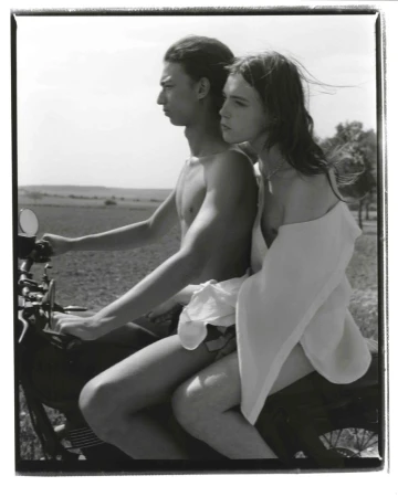 Black and white photograph of a young couple riding a motorcycle through a rural landscape on a sunny day, the woman wearing a white garment.