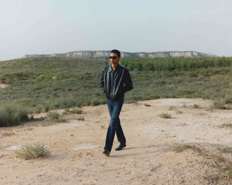 Adult male in sunglasses and a jacket walks across a sandy desert landscape with sparse vegetation on a bright day with a hill in the distance.