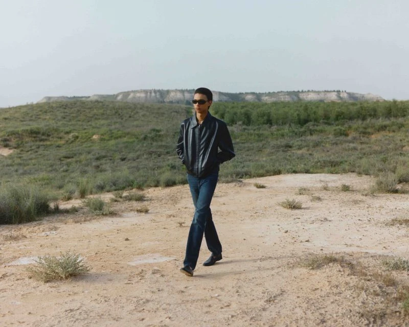 Adult male in sunglasses and a jacket walks across a sandy desert landscape with sparse vegetation on a bright day with a hill in the distance.
