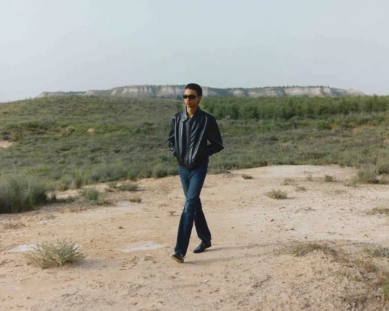 Adult male in sunglasses and a jacket walks across a sandy desert landscape with sparse vegetation on a bright day with a hill in the distance.